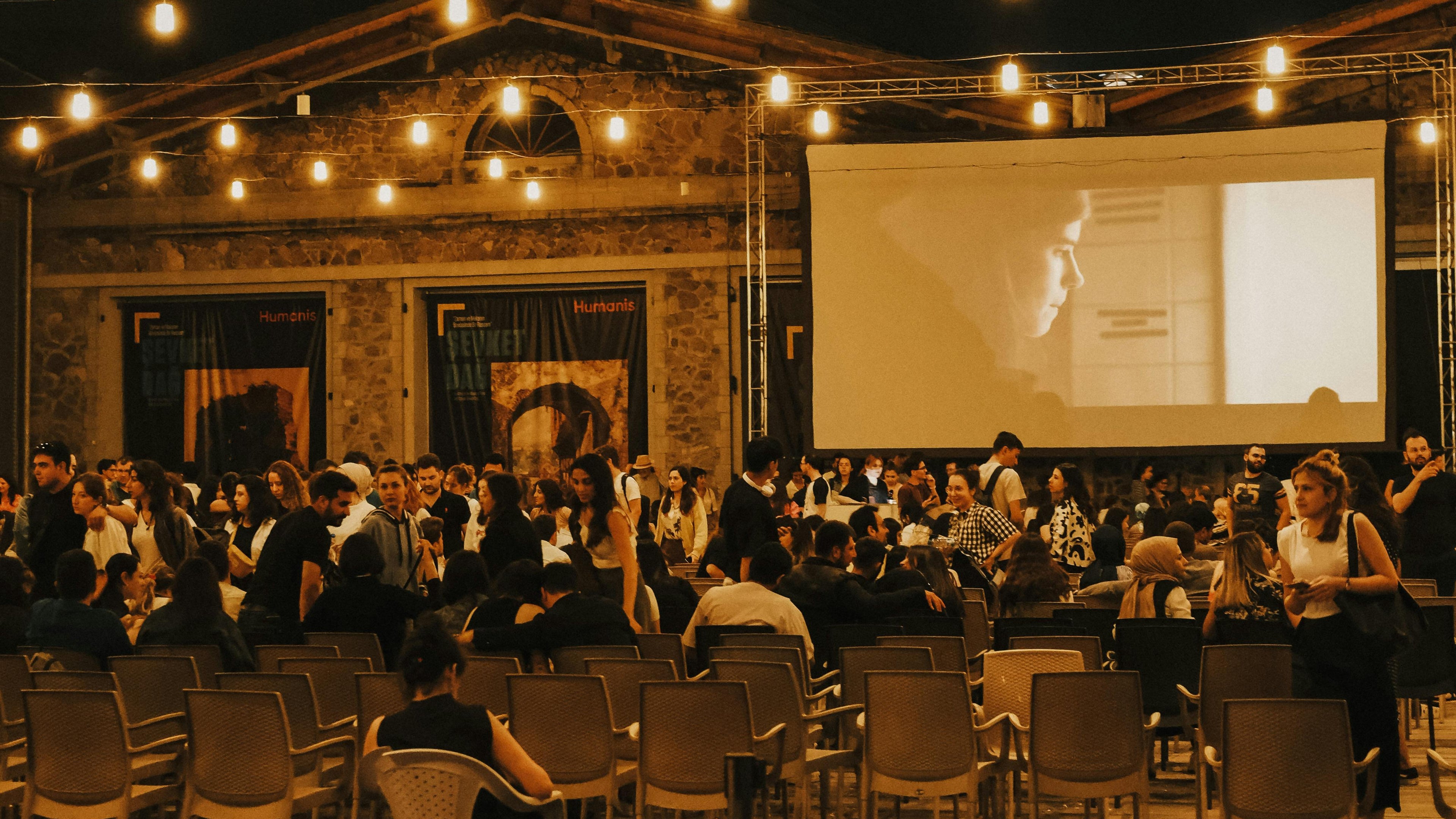 Outdoor event with string lights and a large screen displaying a presentation.